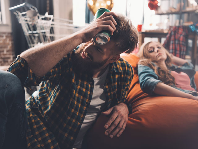 A man holding a cold beer to his head because he has a headache from drinking alcohol